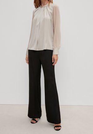 Femme portant un chemisier beige à manches longues plissées et manches transparentes, un pantalon noir à jambes larges et des talons ouverts noirs, debout sur un fond neutre.