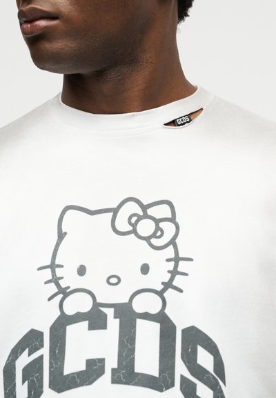 Κοντινή λήψη ενός ατόμου που φοράει λευκό πουκάμισο με μια μικρή σχισμή κοντά στον γιακά και ένα γκρι γραφικό Hello Kitty πάνω από μεγάλα γράμματα "GCDS".