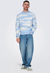 Lyseblå sweater med hvidt sky-mønster, ribbede manchetter, parret med lyseblå løstsiddende jeans og sneakers med grønne accenter.