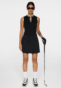 Svart ärmlös golftklänning med dekorativ halsringning, veckad kjol och rak passform. Kompletteras med svarta solglasögon och golfhandske.