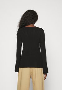 Pull noir côtelé à manches longues, col rond et poignets légèrement évasés ; porté avec un pantalon beige à jambes larges.