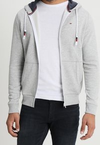 Světlě šedá mikina na zip s tmavě modrou vnitřní podšívkou, šňůrkami a předními kapsami, vrstvená přes bílé tričko, doplněná tmavými džínami.