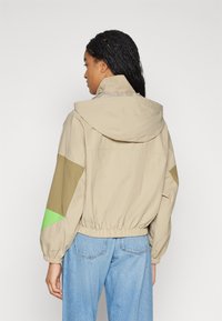 Beige kapuutsiga jope roheliste ja khaki aktsentidega, elastne vöökoht ja laiad varrukad. Tagantvaates on näha tekstureeritud kangast ning sõbralikku lõiget.