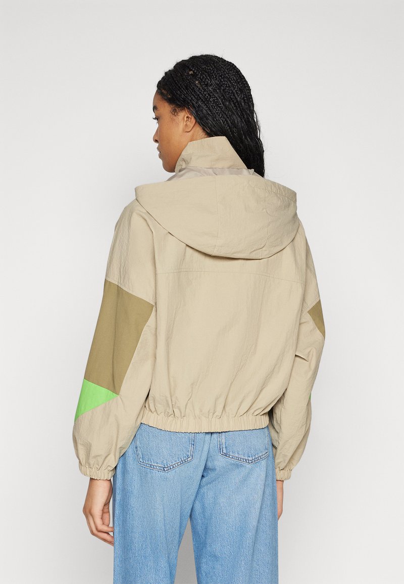 Chaqueta con capucha beige con acentos en verde y caqui, cinturilla elástica y mangas holgadas. La vista trasera muestra una tela texturizada y un ajuste informal.