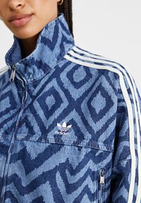 Denim jas met een abstract blauw patroon, hoge kraag en ritssluiting. Bevat witte gestreepte accenten en een klein logo op de borst.