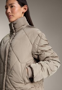 Giubbotto trapuntato beige con alto collo, vestibilità oversize e tasche laterali. Presenta un tessuto liscio con un leggero lucido e cuciture geometriche.