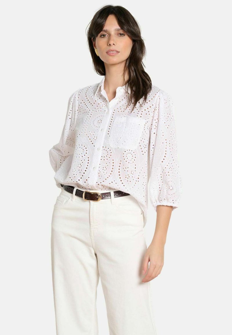 Kobieta ubrana w białą koszulę na guziki z ażurowym wzorem i rękawami do połowy łokcia, włożoną w białe spodnie z wysokim stanem i ciemnobrązowym paskiem.