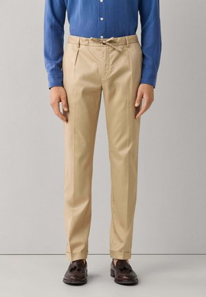 Homme portant un pantalon beige plissé avec taille à cordon, une chemise bleue à boutons et des mocassins à pampilles marron foncé sur un fond uni.
