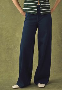 Weite, dunkelblaue Jeanshose mit hoher Taille und lässigem Schnitt, kombiniert mit einem gestreiften, grünen und weißen Hemd.