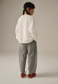 Valkoinen huppari, musta-valkoiset houndstooth-housut, punaiset kengät, joissa on teksturoitu pohja, letitetty hius, kääntynyt pois kamerasta.