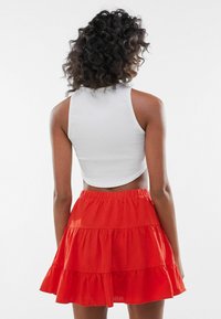 Kvinna med lockigt hår iförd en ärmlös vit crop top och en lager-på-lager röd kjol, stående med ryggen vänd mot kameran.