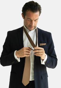 Marineblazer med en hvid skjorte og brunt slips. Slipsen er knyttet i hænderne, med et mønstret lommefyr synligt i jakkens venstre lomme.