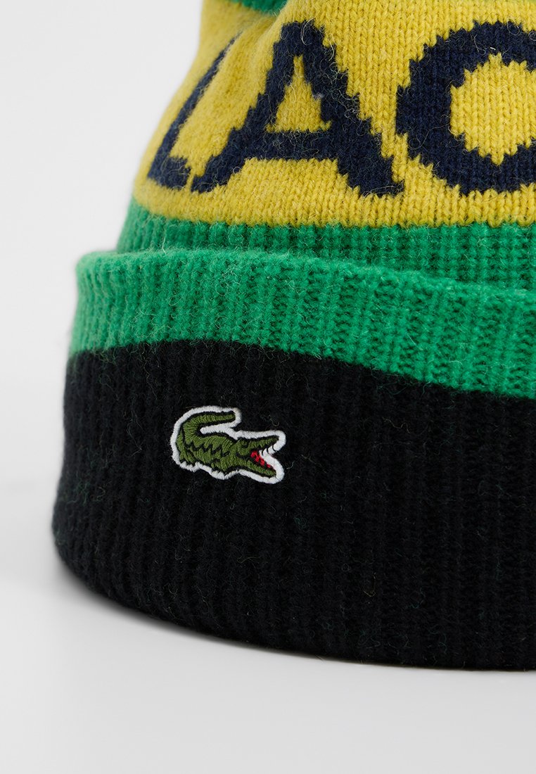 Gebreide muts in geel, groen en zwart. Heeft grote tekst en een klein geborduurd krokodillenlogo op het zwarte gedeelte.