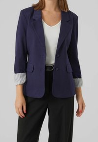Blazer bleu marine avec une coupe ajustée, poches avant et accents de poignets rayés. Porté sur un haut blanc et un pantalon noir. Texture lisse.