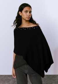 Poncho negro acanalado con dise�ño de hombro al descubierto, que presenta tachuelas metálicas a lo largo del escote. Textura de punto suave, superpuesto sobre una blusa de color gris oscuro.