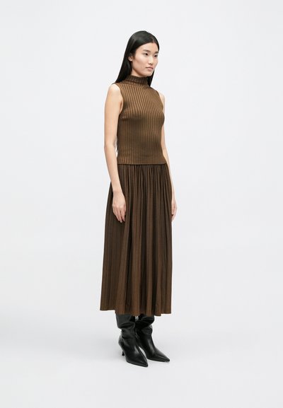 Καφέ ριχτό αμάνικο μπλουζάκι με ψηλό γιακά, συνδυασμένο με μια ρέουσα φούστα σε σκουρότερη απόχρωση του καφέ. Το μοντέλο φοράει μαύρες μπότες με μυτερή μύτη.