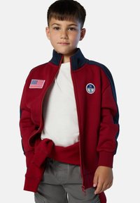Chaqueta roja con cremallera y acentos en color marino, con parche de la bandera americana. Cuenta con un alto cuello, puños acanalados y logo bordado. Textura suave.