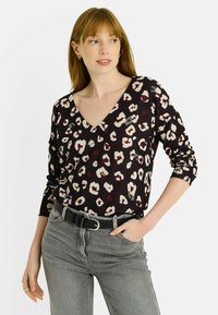 Mujer vistiendo una blusa sin mangas de leopardo en tonos negro y beige con escote en V, jeans grises, cinturón negro, aretes de aro dorados y anillos, posando con una mano en el bolsillo.