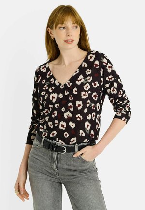 Mujer vistiendo una blusa sin mangas de leopardo en tonos negro y beige con escote en V, jeans grises, cinturón negro, aretes de aro dorados y anillos, posando con una mano en el bolsillo.
