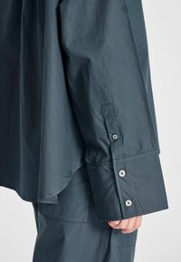 Maglietta a maniche lunghe grigia scura con tre bottoni sul polsino, indossata sopra pantaloni abbinati, mano parzialmente visibile su uno sfondo chiaro.