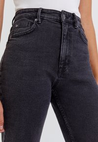Jeans negros de cintura alta con una textura suave, diseño de cinco bolsillos y detalles en tono plateado. Presentan una silueta ajustada y un ligero desgaste.