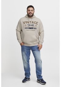 Sudadera beige con texto "VINTAGE Denim", bolsillo canguro, combinada con jeans azules y zapatillas negras, de pie frente a un fondo blanco.