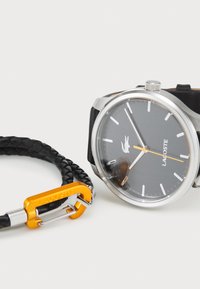 Svart analog klocka med silverfärgad klockkropp, grå urtavla och vita timmarkörer; flätad svart läderarmband med oranga metallspänne.