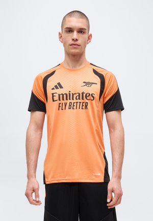 Uomo che indossa una maglia da calcio dell'Arsenal arancione e nera con il logo Emirates Fly Better, in piedi davanti a uno sfondo chiaro e semplice.