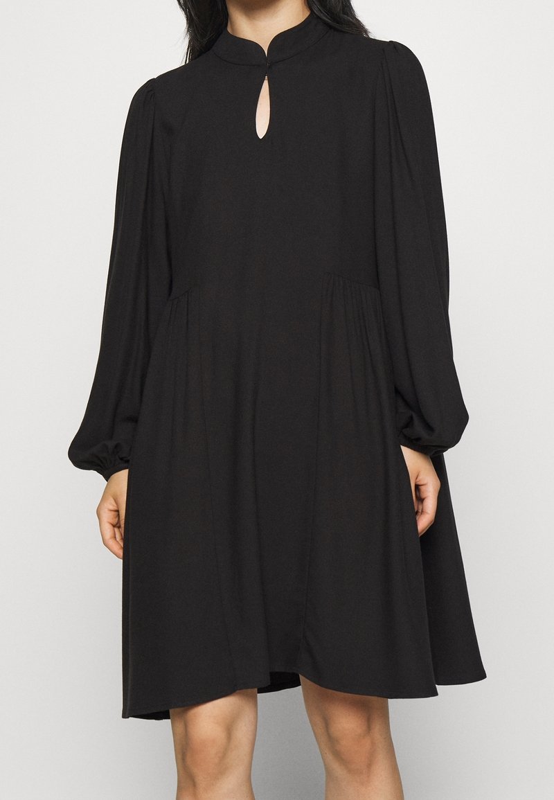 Svart klänning med hög hals, nyckelhålsdetalj och långa puffärmar. Tillverkad av mjukt tyg med en avslappnad, utflugen silhuett och veckade detaljer.