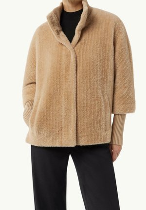 Beige fuzzy kort frakke med bred krave over sort ribstrikket sweater, kombineret med sorte bukser, båret af en stående person med den ene hånd i lommen.