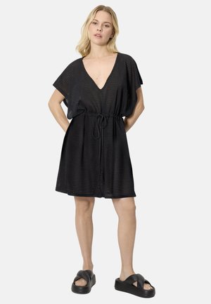 Mujer con un vestido negro de manga corta y cuello en V, con cinturón en la cintura y sandalias plataforma negras, de pie con las manos detrás de la espalda.