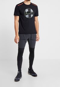 Sort trænings-T-shirt med et cirkulært Nike-logo, parret med mørkegrå træningsbukser med tydelige lapper og mesh-struktur, afsluttet med sorte sko.