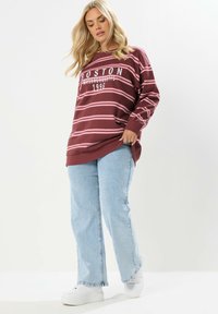 Blondynka w bordo i różowe pasy swetrze z napisem "Boston Massachusetts 1996", jasnoniebieskich dżinsach i białych trampkach na jednolitym tle.