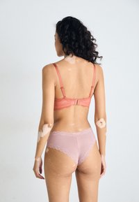 Oransje BH med blonder og rosa truse med bølgete kanter. Myke teksturer står i kontrast til den glatte stoffet, sett bakfra.