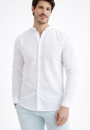 Jeune homme aux cheveux courts et foncés, portant une chemise blanche à boutons et un pantalon bleu clair, debout avec les mains détendues.