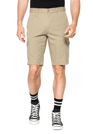 Uomo che indossa pantaloncini cargo beige, sneakers alte nere e calzini neri con strisce bianche, in piedi davanti a uno sfondo bianco.
