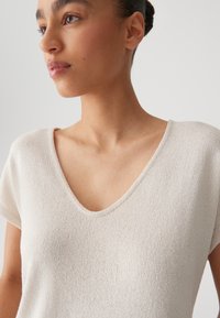 Maglia in tonalità crema chiaro con una finitura testurizzata, scollo a V e maniche corte, caratterizzata da una vestibilità rilassata e un tessuto morbido e traspirante.