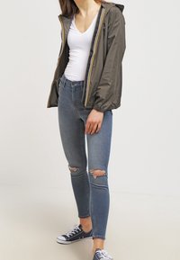 Lekka, oliwkowa kurtka z zamkiem błyskawicznym i kapturem, z elastycznymi mankietami i żółtymi akcentami, w zestawieniu z niebieskimi obcisłymi dżinsami z podartymi kolanami.