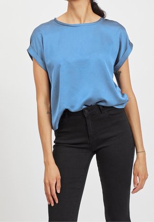 Mujer vestida con una blusa azul de satén de manga corta y suelta, metida dentro de unos vaqueros pitillo negros de cintura alta, de pie contra un fondo liso.