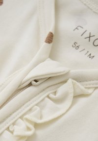 Helles beige Kleidungsstück mit einer weichen Textur, ausgestattet mit einem Reißverschluss und Rüschenakzenten am Ausschnitt. Kleine braune Muster sind sichtbar.