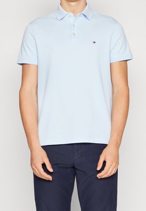 Mężczyzna ubrany w jasnoniebieską koszulkę polo z krótkim rękawem i trzema guzikami oraz granatowe spodnie, stojący na jednolitym tle.