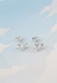 Silverfärgade fjärilspärhängen med intrikata utskurna mönster, mjuka kurvor och polerade ytor, placerade mot en ljusblå bakgrund.