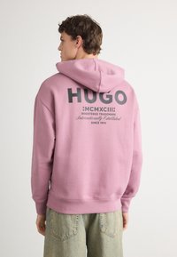 Pulover cu glugă roz, având un mare logo negru "HUGO" pe spate, cu o textură moale și detalii cu tiv căptușit. Asortat cu blugi de culoare albastru deschis.