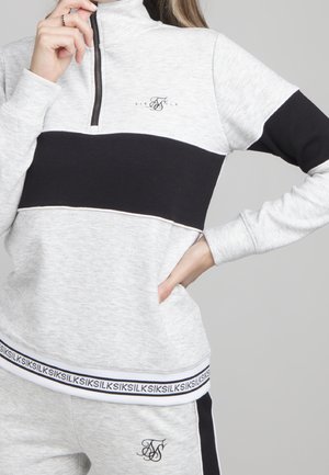 Personne portant un sweat zippé bicolore gris clair et noir et un pantalon assorti avec des logos de marque et des détails textuels.