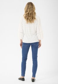Haut à manches longues blanc avec un motif en œillets, manches froncées, associé à un jean skinny bleu foncé et des ballerines noires. Vue arrière de l'ensemble.