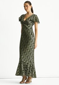Long green dress with a V-neck, ruffled sleeves, and gold speckled pattern. Flared hem with a ruffle detail. Model wearing strappy heels.