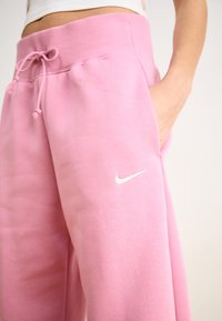 Roze Nike joggingbroek met een geribbelde tailleband, trekkoord en zijzakken. Bevat een wit Nike-logo op de linker bovenbeen. Zachte textuur.