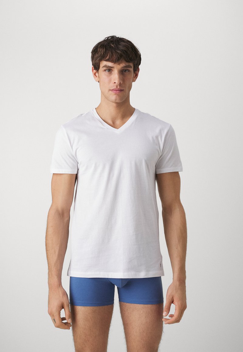 Balti medvilninė V formos kaklo marškinėliai trumpomis rankovėmis, dėvimi virš mėlynų bokserinių šortų. Audinys atrodo lygus, su minimalistiniu dizainu.