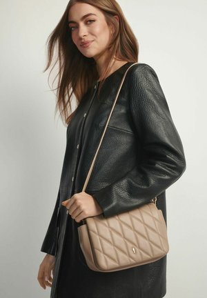 Młoda kobieta w czarnym, teksturowanym płaszczu trzyma beżową pikowaną torbę na ramię na jednolitym tle.