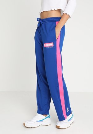 Person wearing blue Reebok Classic track pants with pink side stripes and white sneakers, standing with one hand in pocket.